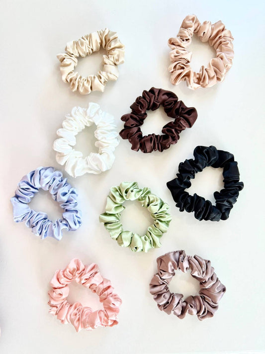 Nine silk scrunchies in assorted colors including beige, pink, white, brown, black, blue, green, and mauve, arranged on a light off-white background.