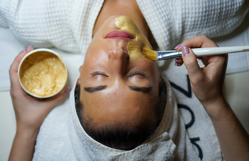 Person applying a facial mask with a brush and container, wrapped in a towel.
